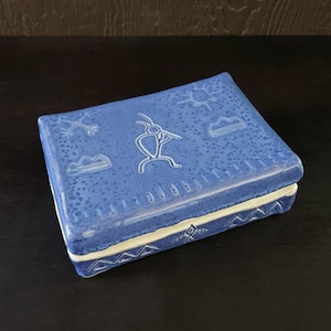May include: A blue ceramic box with white trim and a design of Native American petroglyphs. The box has a textured surface and a white interior.