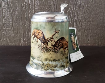 Könnte beinhalten: Ein Steinkrug aus Zinn mit Deckel und Henkel. Der Krug ist mit einer Szene von zwei Hirschen verziert, die sich in einem Wald kämpfen. Der Krug steht auf einer Holzoberfläche.