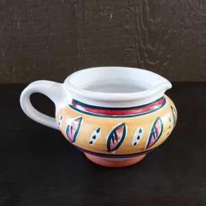 May include: A white ceramic creamer with a handle, decorated with a yellow, orange, red, and green leaf pattern.
