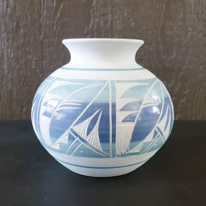 Puede incluir: Un jarrón de cerámica blanca con un diseño geométrico azul y blanco. El jarrón tiene un cuerpo redondeado y una boca ancha.