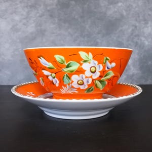 May include: An orange ceramic bowl and saucer set. The bowl and saucer are decorated with white flowers, green leaves, and purple accents. The rim of the saucer has a gold trim. The bowl is sitting on top of the saucer.