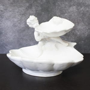 Puede incluir: Un cuenco decorativo de cerámica blanca con una figura de querubín que sostiene dos platos en forma de concha. El querubín está situado entre los dos platos, que están apilados uno encima del otro. El cuenco tiene un borde festoneado.