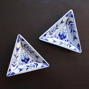 Puede incluir: Dos platos triangulares de cerámica blanca con diseños florales y arquitectónicos azules. Los platos están dispuestos sobre una superficie marrón oscuro. Los diseños azules incluyen flores estilizadas, hojas y motivos de edificios.