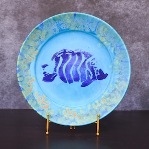 Puede incluir: Un plato redondo de cerámica con un centro azul claro y un diseño de pez azul oscuro. El borde tiene un patrón texturizado en tonos azules y amarillos. El plato se exhibe sobre un soporte de metal dorado.
