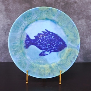 Puede incluir: Un plato redondo de cerámica con un diseño de pez azul. El plato tiene un centro azul claro con una ilustración de pez azul oscuro. El borde es una mezcla de azul claro y verde. El plato se exhibe sobre un soporte dorado.