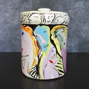 May include: A cylindrical ceramic jar with a lid, featuring a hand-painted design. The jar's exterior showcases colorful, abstract faces in shades of orange, blue, and purple. The lid is white with black line art. The jar is a decorative piece.
