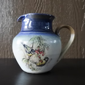May include: A ceramic pitcher with a blue rim and handle. The body of the pitcher is white with a painted design of two birds and red berries. The handle is tan and the pitcher has a spout.