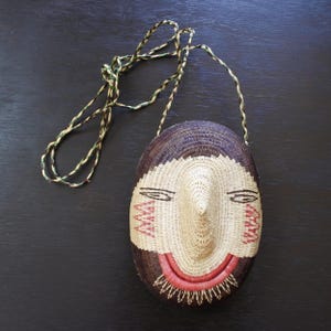 Puede incluir: Bolso ovalado de mimbre tejido con diseño de rostro. El bolso presenta un rostro tejido con cabello marrón oscuro, una cara beige y detalles en rojo y negro. Tiene una correa trenzada de hilos verdes, beige y marrones.