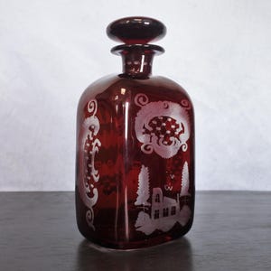 May include: A dark red glass decanter with a square body and a stopper. The decanter is decorated with an etched design of a house, trees, and floral patterns.