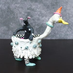 Puede incluir: Una escultura de cerámica caprichosa que presenta un perro negro con lunares blancos dentro de un recipiente blanco y plumoso. El recipiente está sostenido por patas azules con piernas a rayas blancas y negras. Una cabeza blanca con forma de cisne con un gorro de fiesta completa el diseño.