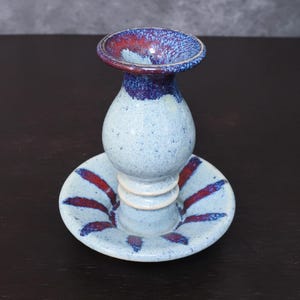 May include: A handmade ceramic candle holder with a light blue speckled finish. The candle holder has a flared top with red and blue accents, a rounded body, and a circular base with red stripes. A unique piece for home decor.