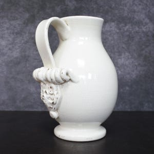 May include: A white ceramic pitcher with a handle and a decorative relief. The pitcher has a bulbous body and a wide base. The handle is curved and attached to the body with a decorative element. The pitcher is approximately 10 inches tall.