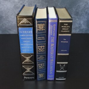 May include: Four vintage hardcover books with gold lettering and decorative designs. Titles include works by O. Henry, Agatha Christie, and Robert Louis Stevenson. The books are in various shades of blue and black.
