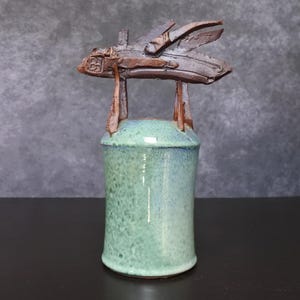 May include: A handcrafted ceramic sculpture featuring a cylindrical base in shades of green and blue. The sculpture is topped with a brown, abstract form supported by vertical brown elements. The artwork has a rustic, textured appearance.
