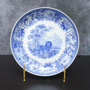 May include: A round ceramic plate featuring a blue and white transferware pattern. The center depicts a lion standing in a landscape with trees and mountains. The wide border is decorated with intricate floral motifs and scrolling foliage.