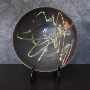 May include: A dark ceramic bowl featuring abstract green and copper-coloured glaze designs. The round bowl is displayed on a black metal stand, highlighting the unique artistic patterns. The bowl's interior has a dark, matte finish.