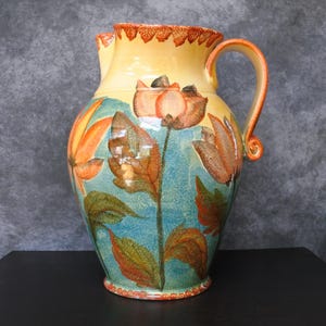Puede incluir: Una jarra de cerámica pintada a mano con la parte superior y el asa amarillas, con diseños florales naranjas y marrones sobre un fondo azul. La jarra tiene una base ancha y un asa curva, perfecta para servir bebidas.