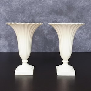 May include: A pair of off-white ceramic vases featuring a fluted, trumpet-shaped body with a scalloped rim. Each vase stands on a square pedestal base with a rounded neck, showcasing a vertical ribbed texture that extends from the base to the top.