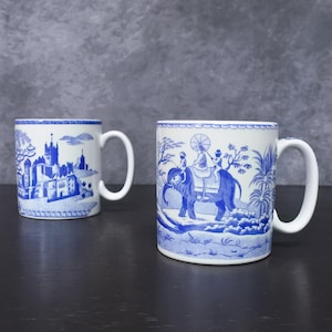 May include: Two white ceramic mugs featuring blue transferware patterns. The left mug displays a detailed castle landscape, while the right mug depicts a person riding an elephant under a parasol surrounded by palm trees and foliage.