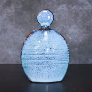 May include: A ceramic decanter with a round body and a small, round stopper. The decanter is light blue with darker blue and brown accents. Horizontal lines encircle the body. The stopper is also light blue with a brown rim.