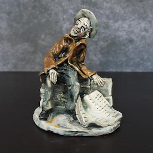 May include: A ceramic figurine of a seated man in a brown jacket and hat, resting on a stone structure. The figure has a weary expression and is next to a scroll. The base is a light color.