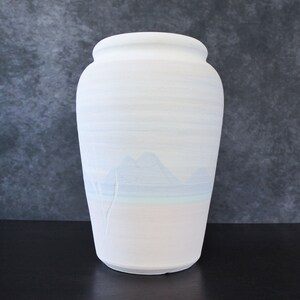 May include: A white ceramic vase with a wide body and a slightly flared rim. The vase features a landscape design with mountains and a tree, painted in shades of blue and white. The vase is approximately 12 inches tall.