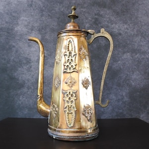 Puede incluir: Una tetera de metal de color dorado y cobre con intrincados diseños florales. La tetera tiene un cuerpo alto y delgado y un pico curvo. El asa está unida a la parte superior del cuerpo y se curva hacia arriba.