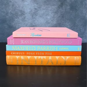 May include: A stack of five hardcover books with colorful spines. The top book is light pink with the text Populuxe. Below it is a magenta book titled Jean Howard's Hollywood, a light blue book titled Maxfield Parrish, and two orange books.