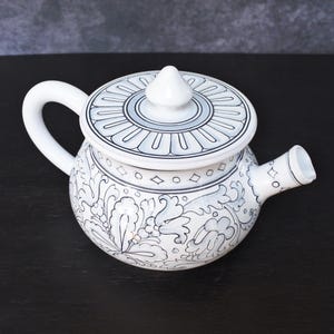 Könnte beinhalten: Weiße Keramik-Teekanne mit Deckel, Henkel und Ausguss. Die Teekanne ist mit schwarzen und grauen floralen und geometrischen Mustern verziert. Der Deckel hat einen zentralen Knopf und einen dekorativen Rand. Zum Aufbrühen von Tee.