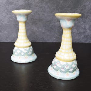 May include: Two ceramic candle holders. The candle holders have a yellow and white checkered pattern on the stem, with a light blue base and top. The base has a scalloped design in green and pink.