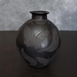 Puede incluir: Un jarrón redondo de color marrón oscuro con un cuello estrecho. El jarrón presenta diseños grabados de mariposas y hojas alrededor de su cuerpo. El jarrón está hecho de un material mate y se encuentra sobre una superficie oscura.