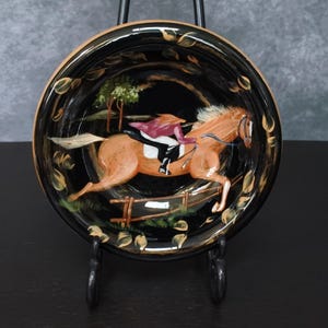 May include: A round, black decorative plate featuring a horse and rider jumping over a fence. The horse is brown with a white mane and tail. The rider wears a maroon jacket and black pants. The plate has a gold leaf design around the rim.