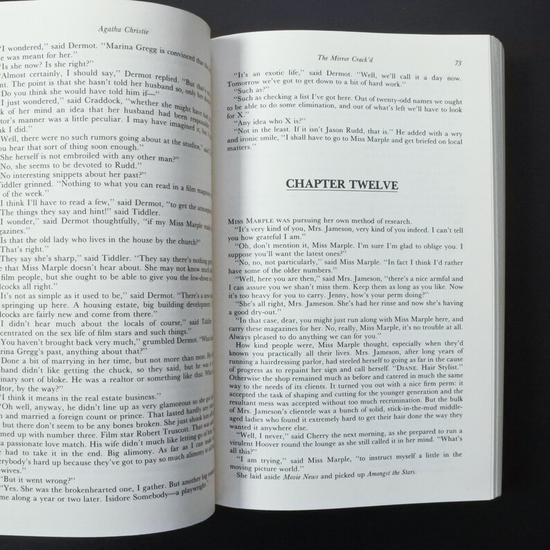 May include: Open book with text from Agatha Christie's "The Mirror Crack'd". The pages are yellowed, and the text is in black. Chapter Twelve is visible. The book is open on a dark surface.