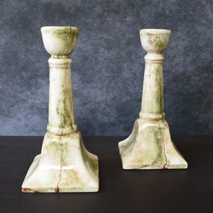 May include: Two white ceramic candle holders with a green and brown marbled design. The candle holders have a square base and a tall, cylindrical column.