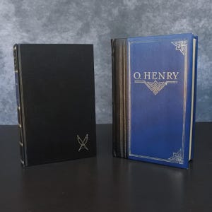 May include: Two hardcover books. One is black with a silver emblem and gold-edged pages. The other has a blue cover with gold lettering that reads "O. HENRY" and decorative gold accents.