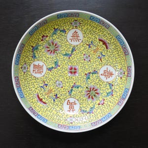 May include: A round, yellow ceramic plate with a detailed floral design. The plate features red, pink, and blue floral patterns, with Chinese characters in circular medallions. The rim has a blue and white geometric border.