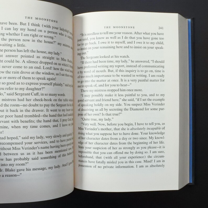May include: Open book with text from "The Moonstone" by Wilkie Collins. The pages are yellowed, and the text is in black. The book is open to a chapter with dialogue. The title and page number are visible.