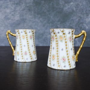 以下が含まれることがあります： 金色の取っ手と花柄が特徴の白い磁器マグカップ2つ。ピンク、黄色、オレンジ色のバラが繰り返し描かれ、金色のトリムと竹風の取っ手がアクセントになっています。クラシックなデザインです。