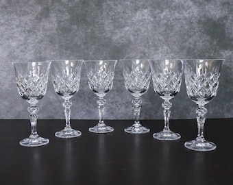 May include: Six clear crystal wine glasses with diamond-cut patterns. The glasses have a classic design with a stemmed base and a wide bowl. The glasses are arranged in a row on a dark surface, with a gray background.