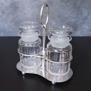 Op de afbeelding: Zilverkleurige kruidenhouder met twee heldere glazen potten met matglazen deksels. De houder heeft een ringvormig handvat en staat op een voet met drie poten.