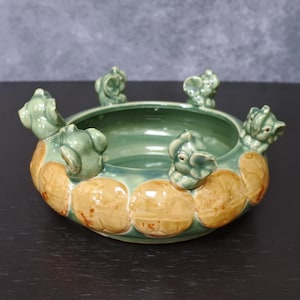 May include: A ceramic bowl with a green interior and exterior, adorned with six elephant figurines around the rim. The bowl features a light brown and tan textured pattern around the base.