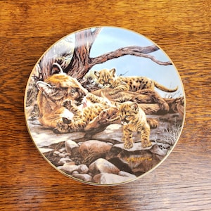 May include: A ceramic plate with a colorful illustration of a mother cougar and her three cubs. The plate has a gold rim and the artist's signature "G. Beecham" is visible in the lower right corner.