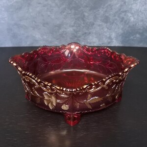 May include: A vintage, ruby-red glass bowl with a decorative, scalloped edge and three feet. The bowl has a floral and leaf pattern with gold detailing. The interior is smooth, while the exterior has a textured design. The bowl is approximately 20 cm in diameter.