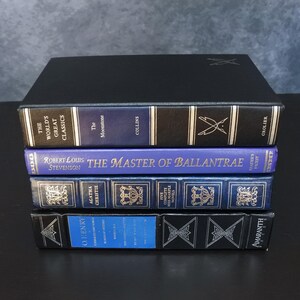 May include: A stack of hardcover books with blue and black spines. Titles include "The World's Great Classics," "The Moonstone," and "The Master of Ballantrae." The books have gold lettering and decorative elements.