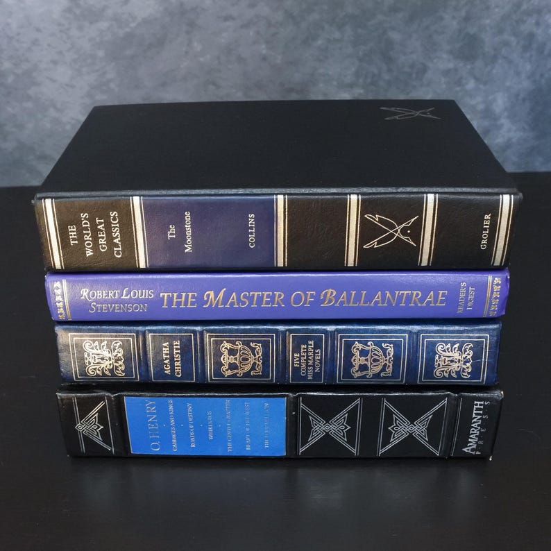May include: A stack of hardcover books with blue and black spines. Titles include "The World's Great Classics," "The Moonstone," and "The Master of Ballantrae." The books have gold lettering and decorative elements.