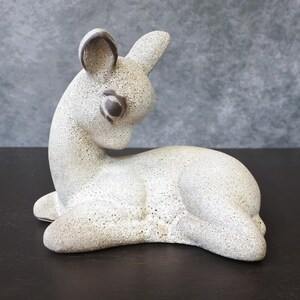 Può includere: Una statuetta di ceramica bianca di un cerbiatto con occhi e orecchie marroni. Il cerbiatto è sdraiato con la testa girata di lato.