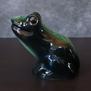 Può includere: Una statuetta di rana in ceramica con una smaltatura nera e verde lucida. La rana è seduta con la testa sollevata e gli occhi chiusi.