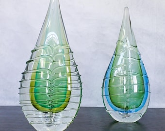 May include: Two teardrop-shaped glass vases with a layered design. The larger vase is clear with green and yellow stripes. The smaller vase is clear with green, blue, and yellow stripes.
