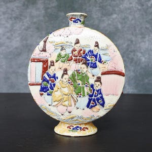 May include: A ceramic vase with a round body and a narrow neck. The vase features a detailed, colorful relief scene of figures in traditional clothing. The base is decorated with floral patterns, and the overall design is ornate and detailed.