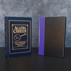 May include: Two hardcover books. The book on the left is dark blue with gold lettering that reads "Agatha Christie" and "Five Complete Miss Marple Novels." The book on the right is black with a purple spine.